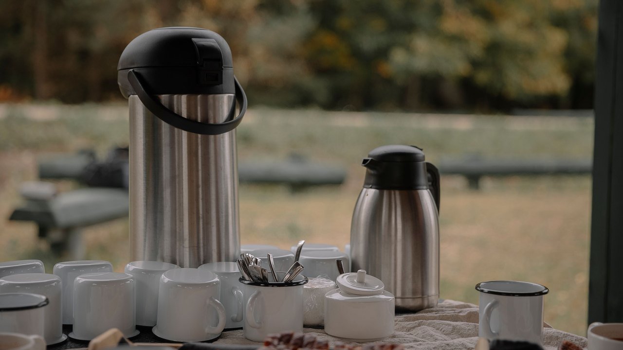 Self-serve drink station with a coffee airpot, thermal jug, rows of white mugs and a tray of cake