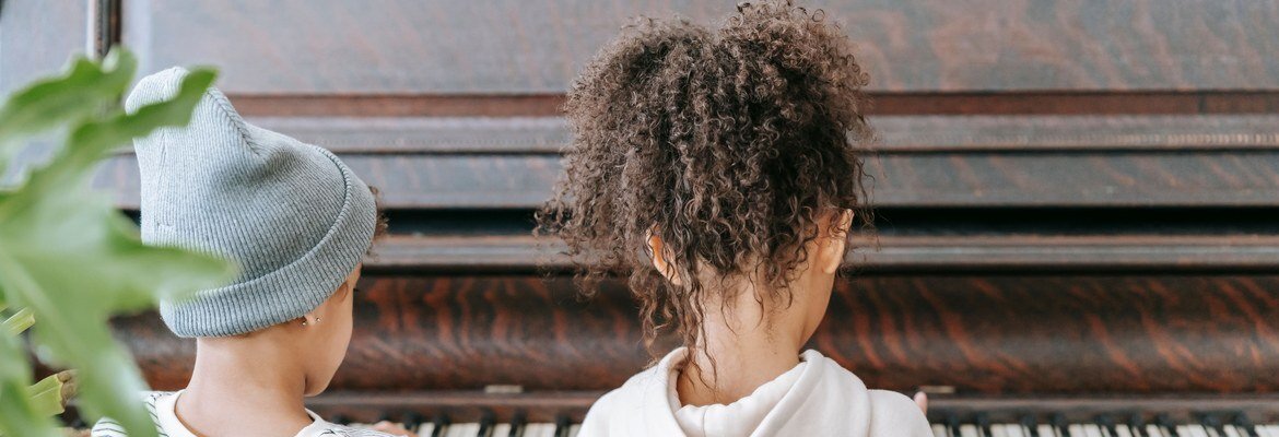 Two young children playing an old wooden upright piano at home, seen from behind