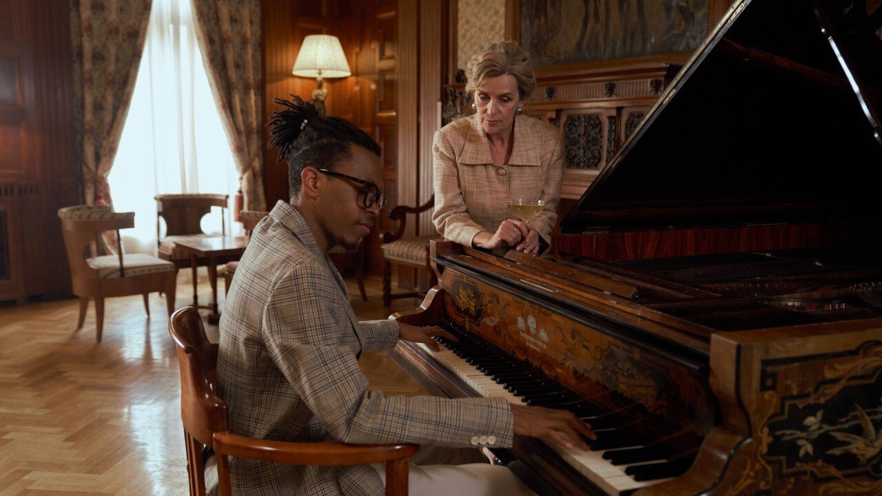 Older piano teacher leaning on a grand piano, watching an adult student's hands as he plays