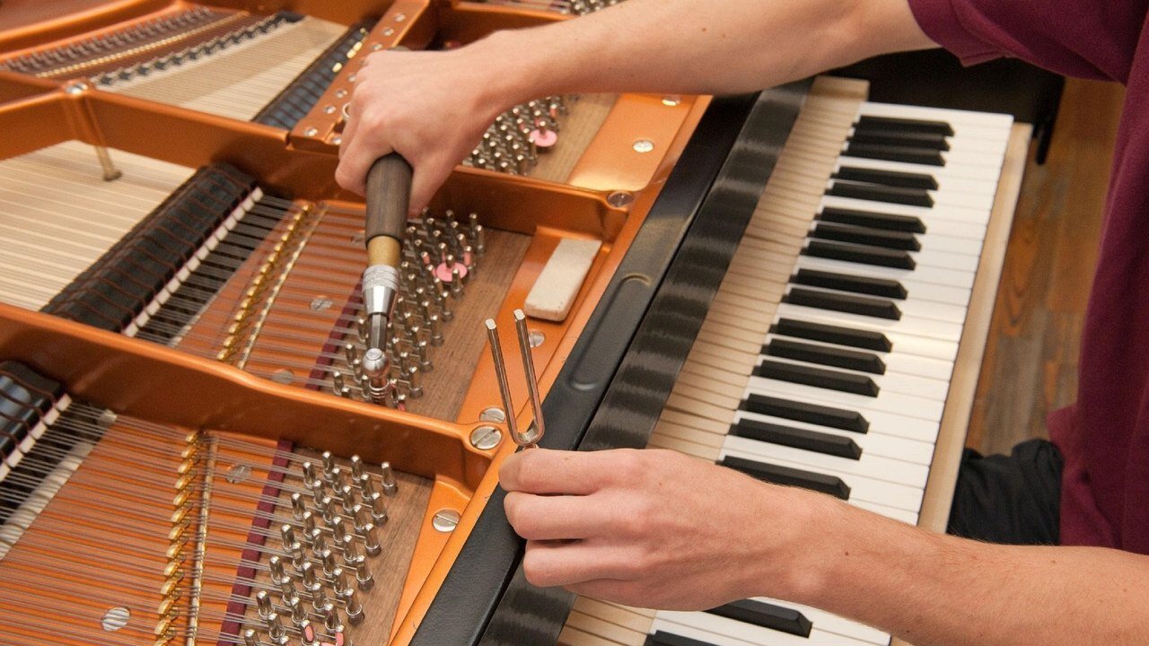 Piano tuner turning a tuning pin with a tuning hammer while holding a tuning fork over the keys