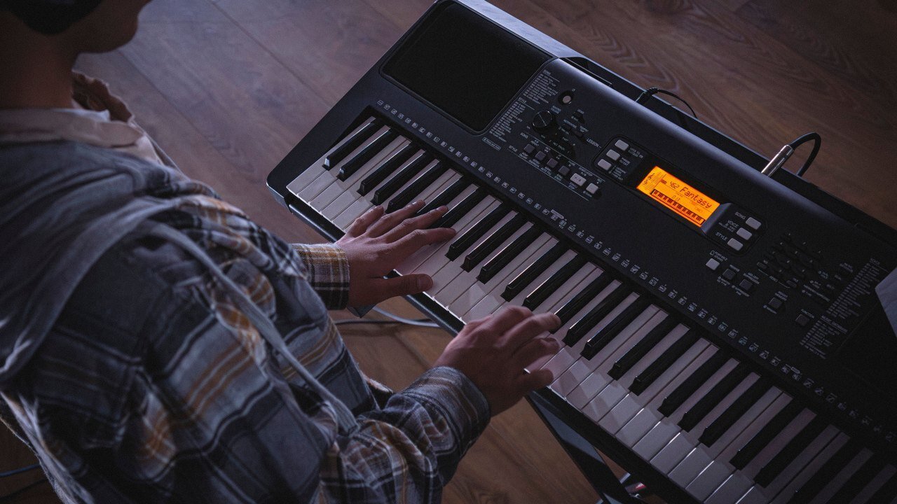 Hands playing an arranger keyboard with an orange screen and style buttons, on a stand over a wood floor