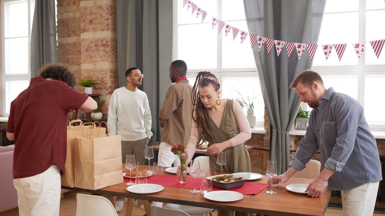 Hosts setting plates, glasses and a dish along one long table in a bright hall before a reception