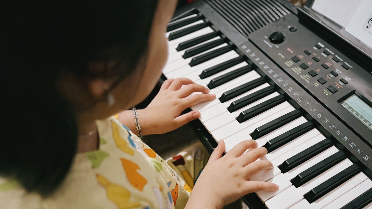 A young child plays an electronic keyboard with curved fingers on the keys, the corner of a lesson page on the stand